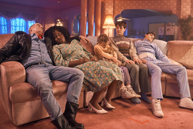 Pięć osób siedzi zgarbionych i nieprzytomnych na różowej kanapie w salonie w stylu retro, ubranych w ubrania vintage. Małe dziecko siedzi na kolanach jednej osoby, a nastrój jest niesamowity i tajemniczy.