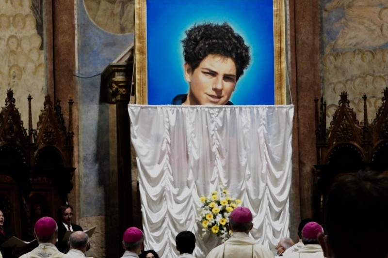 Duży portret młodego mężczyzny z kręconymi włosami jest eksponowany nad ołtarzem okrytym białym płótnem, z żółtymi kwiatami z przodu. Kilku duchownych w białych szatach i różowych czapkach stoi naprzeciwko portretu w kościele.