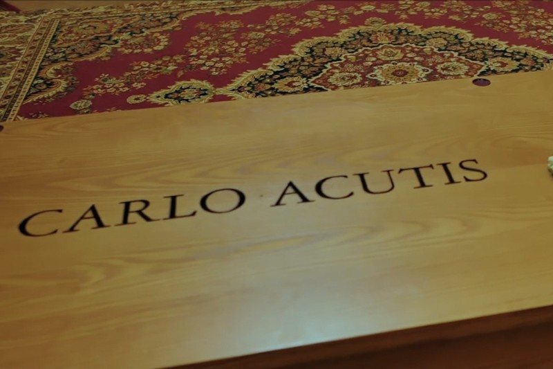 Drewniana powierzchnia z wygrawerowanym napisem CARLO ACUTIS, położona na wzorzystym dywanie w kolorze czerwonym i złotym.