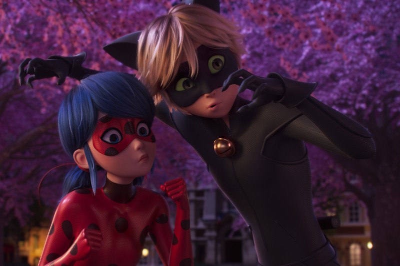 Dwie animowane postacie, Biedronka i Czarny Kot, w kostiumach superbohaterów, stoją blisko siebie, wykonując figlarne, kocie gesty pazurów. W tle widać różowo kwitnące drzewa i budynki.