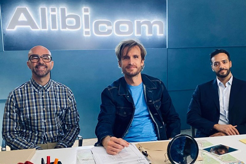 Trzech mężczyzn siedzi przy biurku z artykułami biurowymi i papierami, pozując i uśmiechając się do kamery. Za nimi jest świecący napis Alibi.com na niebieskiej ścianie.