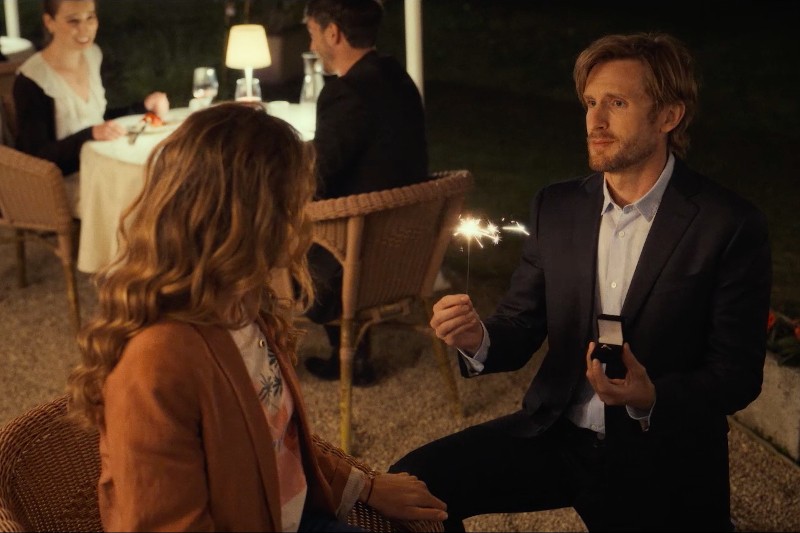 Mężczyzna w garniturze klęka i oświadcza się kobiecie w restauracji na świeżym powietrzu, trzymając w jednej ręce pudełko na pierścionek, a w drugiej zapaloną zimną wodę. W tle widać innych gości.