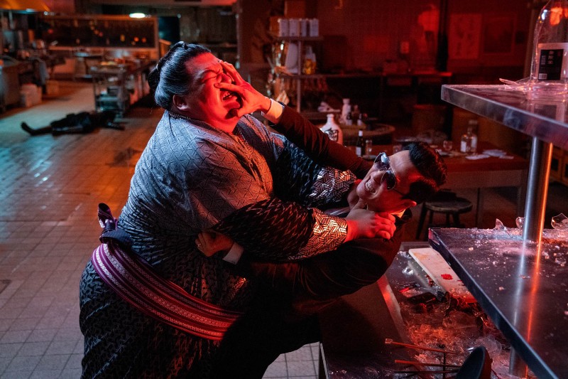 Podczas kłótni w restauracji mężczyzna we wzorzystym szlafroku łapie za twarz mężczyznę w garniturze. W tle widać rozbite szkło na ladzie i postać leżącą na podłodze.