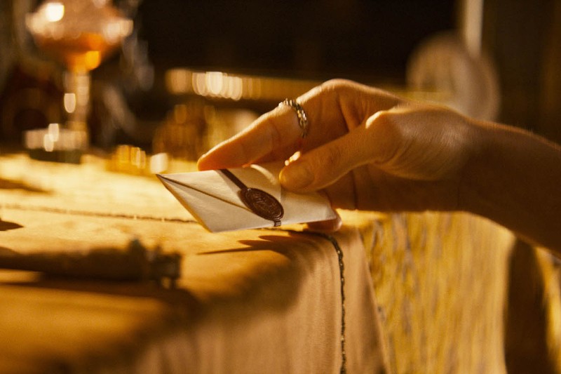 Dłoń z pierścieniami kładzie małą, zapieczętowaną kopertę z czerwoną pieczęcią woskową na stole przykrytym złotym obrusem. Tło jest delikatnie rozmyte, co daje ciepłą i intymną atmosferę.