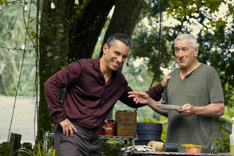 Dwóch mężczyzn stoi na zewnątrz przy stole. Mężczyzna po lewej uśmiecha się i pochyla do przodu, trzymając rękę na ramieniu drugiego. Starszy mężczyzna po prawej trzyma talerz i wygląda na zrelaksowanego. W tle widać drzewa i lampki choinkowe.