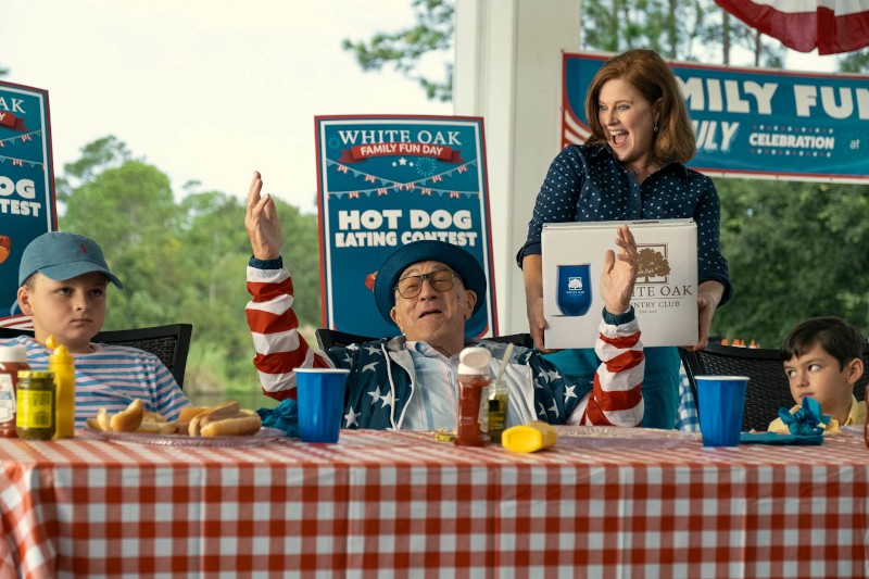 Mężczyzna w kurtce w gwiazdy i paski świętuje konkurs jedzenia hot dogów, unosząc ręce, podczas gdy dwóch chłopców siedzi obok niego przy stole w kratkę. Uśmiechnięta kobieta stoi za nimi, trzymając pudełko z nagrodami. W tle widać świąteczne znaki.
