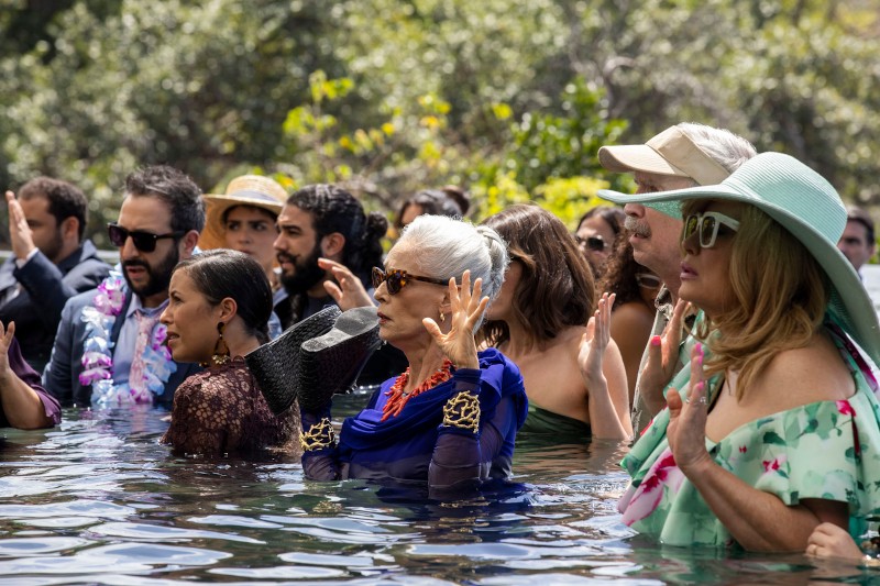 Grupa osób ubranych w kolorowe ubrania i kapelusze stoi po pas w wodzie z uniesionymi rękami, uczestnicząc w ceremonii lub wydarzeniu odbywającym się na świeżym powietrzu, wśród zieleni.