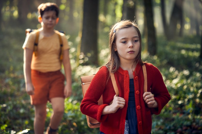 Młoda dziewczyna z plecakiem idzie przez rozświetlony słońcem las, wyglądając na zamyśloną. Chłopiec z plecakiem podąża za nią. Oboje dzieci są otoczeni wysokimi drzewami i rozproszonym światłem słonecznym.