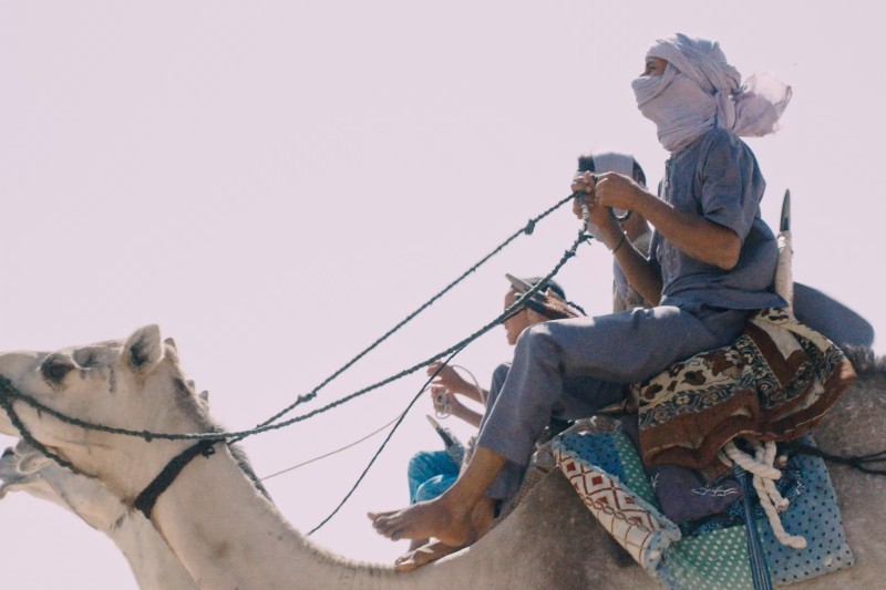 Dwie osoby w chustach na głowach jadą na wielbłądach ozdobionych kolorowymi kocami i siodłami. Jeźdźcy trzymają lejce, a scena jest kręcona z niskiego kąta na tle jasnego, bladego nieba.