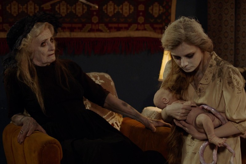 Dwie kobiety o bladych twarzach i ciemnym makijażu siedzą w ciemnym pokoju; jedna ubrana jest na czarno na krześle, druga w beżu, trzymając realistyczną lalkę. Scena ma ponurą, niesamowitą atmosferę.