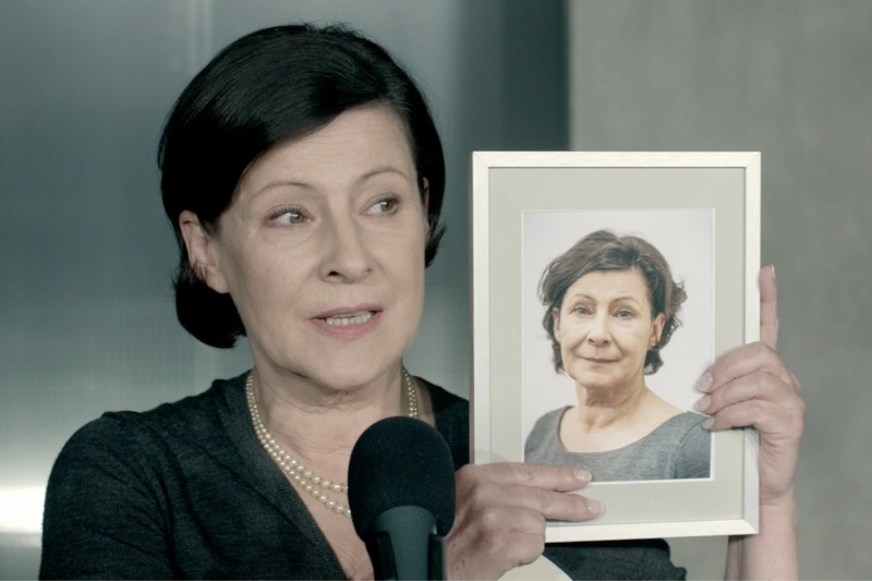 Kobieta z krótkimi, ciemnymi włosami trzyma oprawione zdjęcie siebie, mówiąc do mikrofonu. Ma na sobie ciemny top i perłowy naszyjnik, a tło jest neutralne i delikatnie oświetlone.