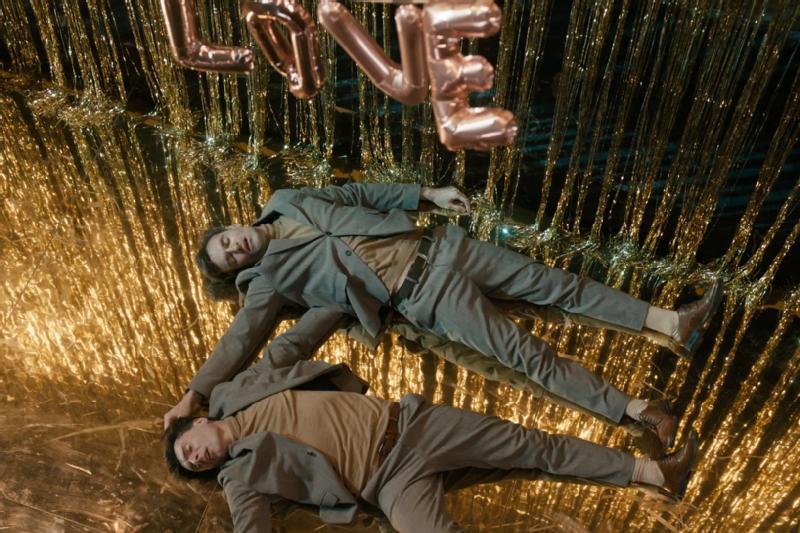 Dwie osoby w pasujących do siebie garniturach leżą obok siebie na błyszczącej złotej powierzchni, trzymając się za ręce. Nad nimi wiszą metalowe litery balonowe, które tworzą napis LOVE. Tło zdobią złote serpentyny.