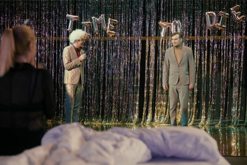 Dwóch mężczyzn w garniturach stoi na scenie z błyszczącym tłem z łańcuchów i złotymi balonami z napisem TIME TO DIE. Na pierwszym planie znajduje się kobieta o blond włosach, a z przodu widać białe łóżko lub koc.