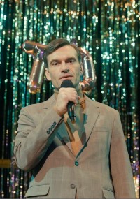 Mężczyzna w beżowym garniturze trzyma mikrofon, stoi przed metalowymi, błyszczącymi serpentynami i srebrnym balonem, sprawiając wrażenie, jakby przemawiał lub śpiewał na scenie.