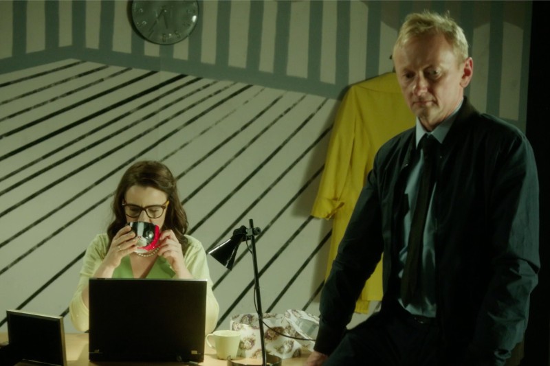 Kobieta siedzi przy biurku z laptopem, pijąc z kubka, podczas gdy mężczyzna w ciemnym garniturze stoi w pobliżu. W tle widać czarno-białe pasy na ścianach, zegar i wiszący żółty płaszcz.