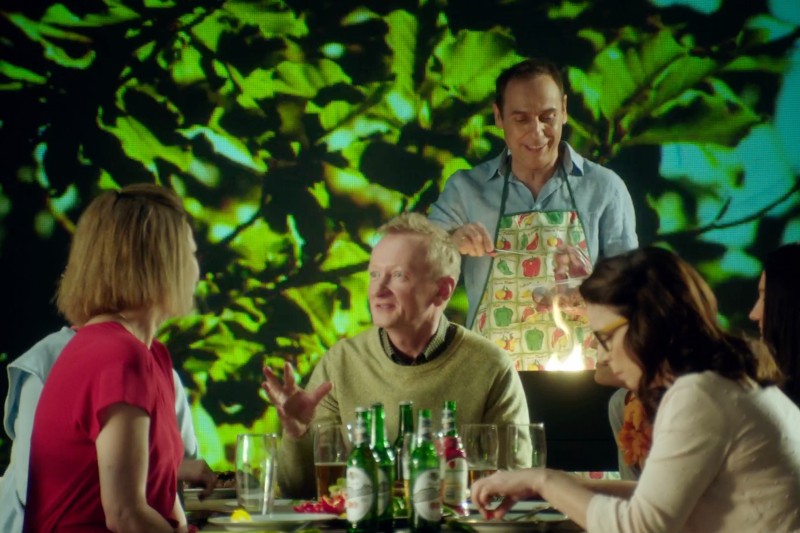 Grupa ludzi siedzi przy stole z jedzeniem i napojami, a mężczyzna w kolorowym fartuchu stoi za nimi i się uśmiecha. Zielone liście są widoczne na jasnym tle. Grupa wydaje się wspólnie cieszyć się ożywionym posiłkiem.