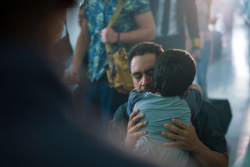 Mężczyzna z zamkniętymi oczami mocno przytula dziecko w zatłoczonym pomieszczeniu, oboje wydają się być wzruszeni. W tle chodzą ludzie z torbami i rozmazanymi postaciami.