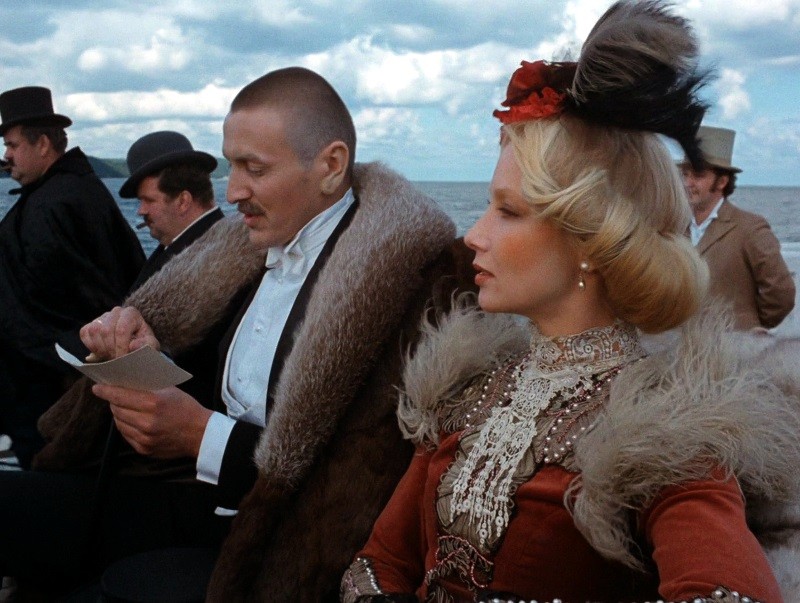 Mężczyzna w futrze pisze na papierze obok kobiety w ozdobnej sukni i kapeluszu z piórami; oboje wyglądają na ubranych w stroje z XIX wieku, siedzą na zewnątrz, a w tle widać morze i zachmurzone niebo.