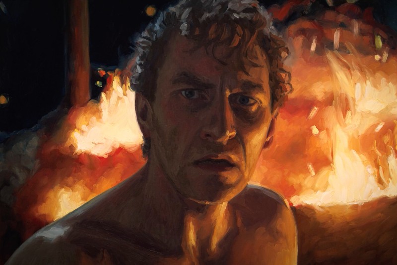 Mężczyzna z nagim torsem i kręconymi włosami stoi przed intensywnymi płomieniami. Wygląda na zmartwionego i oświetlonego blaskiem ognia w dramatycznej nocnej scenie.