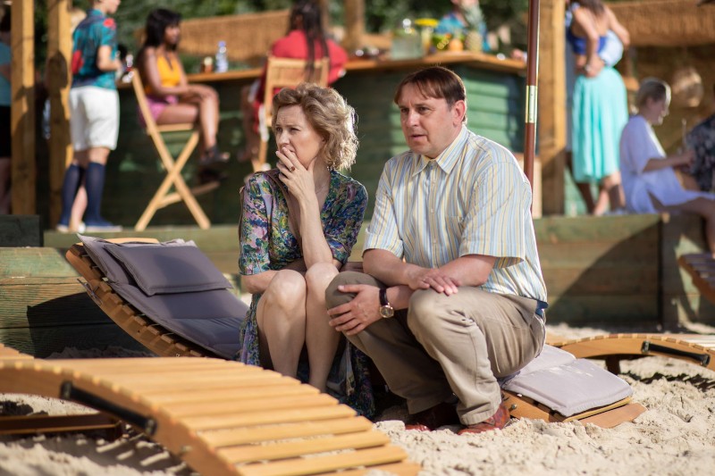 Kobieta i mężczyzna siedzą blisko siebie na leżaku na piaszczystej plaży, wyglądając poważnie i zamyśleni. Inni ludzie towarzysko rozmawiają w tle przy drewnianych stołach i krzesłach.