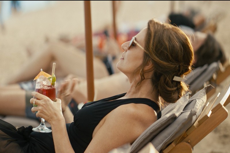 Kobieta w okularach przeciwsłonecznych i czarnym kostiumie kąpielowym relaksuje się na leżaku na plaży, trzymając w dłoni czerwony koktajl z lodem i dodatkami, podczas gdy inni wylegują się w tle na piaszczystej plaży.