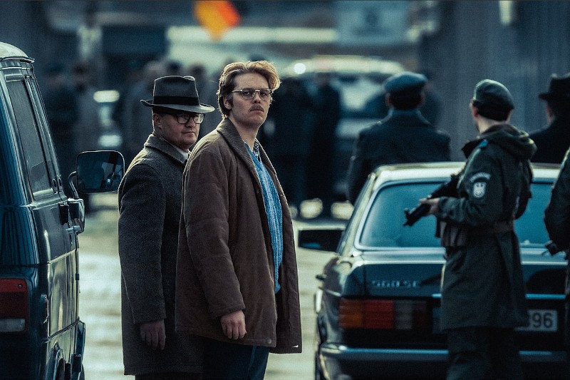 Dwóch mężczyzn w ubraniach vintage stoi na ulicy, patrząc wstecz z poważnymi minami. W pobliżu znajdują się uzbrojeni strażnicy i policyjne pojazdy, co sugeruje napiętą lub dramatyczną sytuację w historycznym otoczeniu.