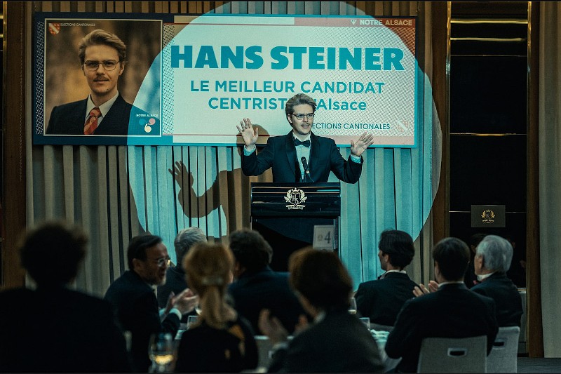 Mężczyzna w garniturze stoi na podium i wygłasza przemówienie do publiczności. Za nim wisi plakat wyborczy z jego zdjęciem i tekstem po francusku, identyfikujący go jako Hansa Steinera, kandydata centrowego w Alzacji.