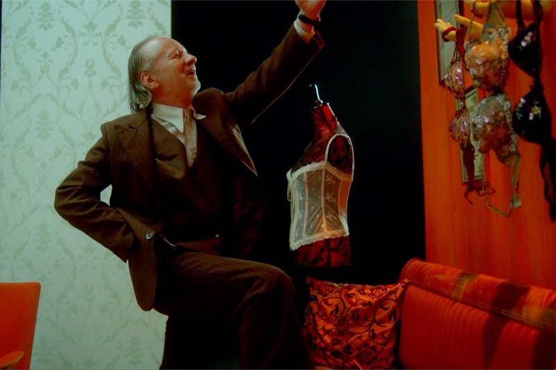 Mężczyzna w brązowym garniturze energicznie pozuje z jedną nogą uniesioną i skierowaną ku górze, w pokoju udekorowanym wzorzystą tapetą, wiszącymi biustonoszami, czerwonym manekinem w bieliźnie i jaskrawymi meblami.