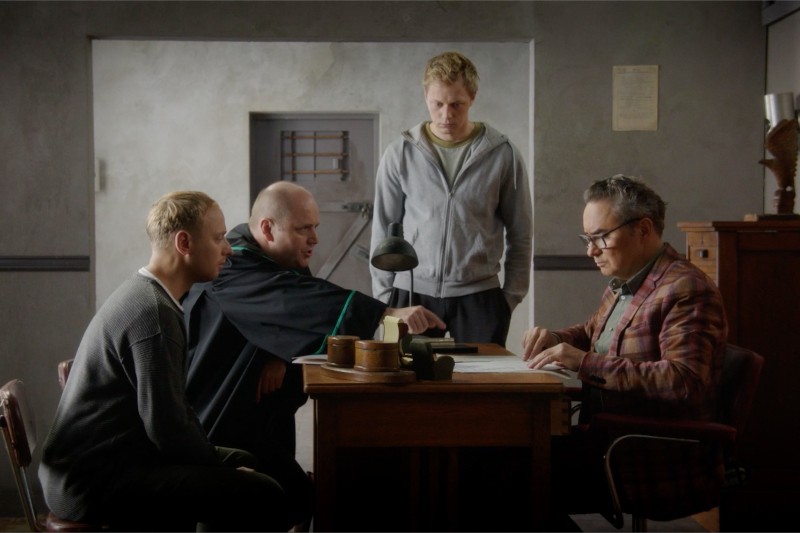 Czterech mężczyzn zgromadziło się wokół drewnianego biurka w słabo oświetlonym pokoju. Dwóch siedzi po jednej stronie, jeden gestykuluje w stronę dokumentów, podczas gdy trzeci stoi za nimi, a inny mężczyzna siedzi naprzeciwko, czytając dokumenty.