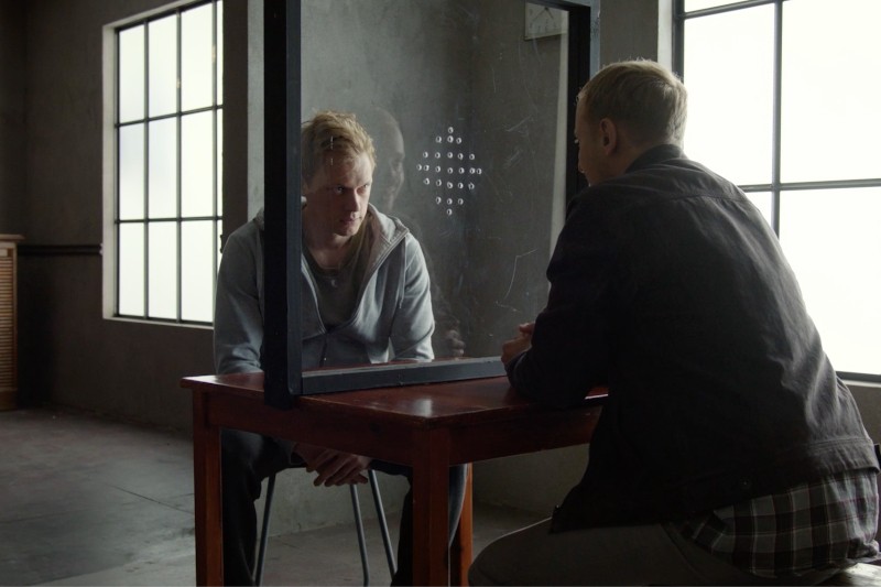 Dwóch mężczyzn siedzi po przeciwnych stronach szklanej przegrody przy stole do odwiedzin w słabo oświetlonym pomieszczeniu. Jeden mężczyzna patrzy w dół, a drugi stoi twarzą do niego, co sugeruje poważną rozmowę.