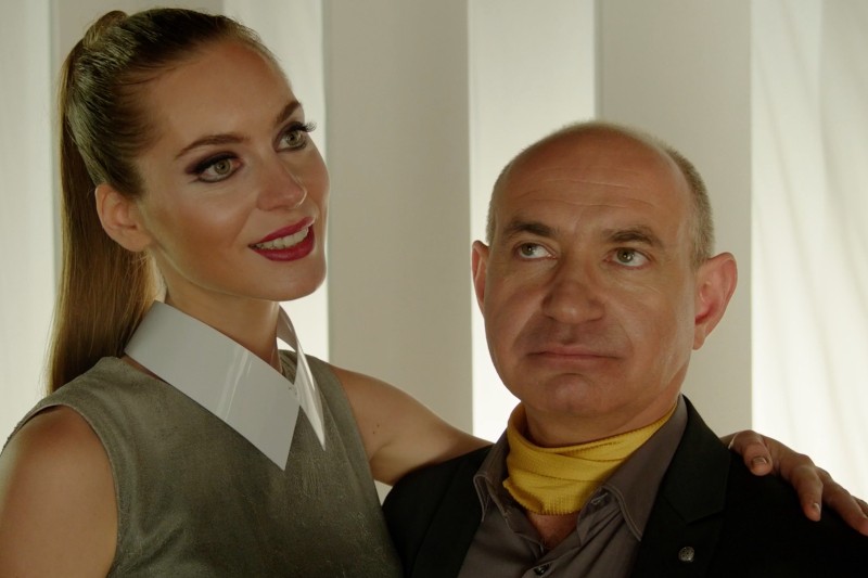 Uśmiechnięta kobieta o długich brązowych włosach i wyrazistym makijażu stoi obok łysego mężczyzny w garniturze z żółtym szalikiem, obejmując go ramieniem. Oboje pozują na tle białych pionowych paneli.