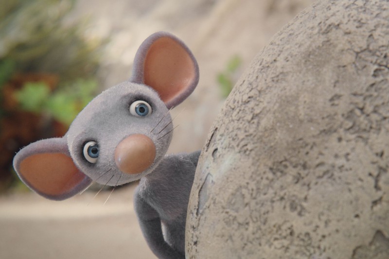 Szerokooka animowana szara mysz z dużymi różowymi uszami wychyla się zza teksturowanego, okrągłego kamienia, wyglądając ciekawie i zaskoczona. W tle widać rozmytą zieleń.