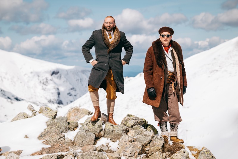 Dwóch mężczyzn w zimowych ubraniach vintage stoi na skalistym terenie z pokrytymi śniegiem górami w tle. Jeden pewnie stoi na skale, podczas gdy drugi stoi obok niego, obaj ubrani w futrzane płaszcze i buty.