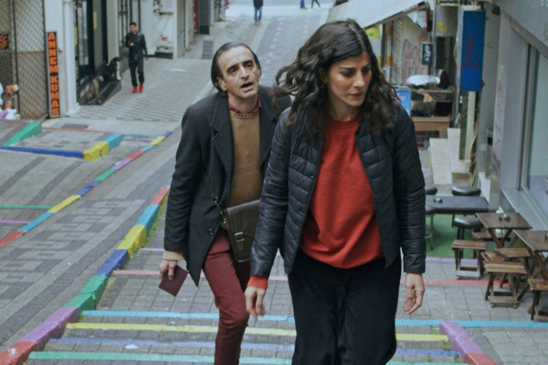 Mężczyzna i kobieta wchodzą po kolorowych, pomalowanych schodach w miejskiej przestrzeni. Mężczyzna ubrany w płaszcz i czerwone spodnie patrzy w górę, podczas gdy kobieta w czarnej kurtce i czerwonym swetrze idzie przed nimi. W tle widać sklepy i stoły.