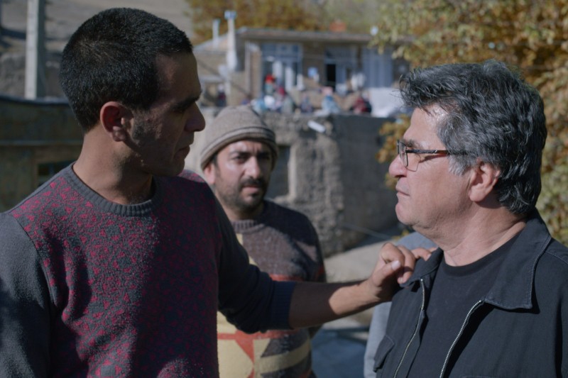 Trzech mężczyzn jest na zewnątrz, jeden z nich kładzie rękę na ramieniu drugiego, prowadząc pozornie poważną rozmowę. Trzeci mężczyzna stoi w tle. Sceneria wydaje się być wiejską okolicą z budynkami i jesiennymi drzewami.