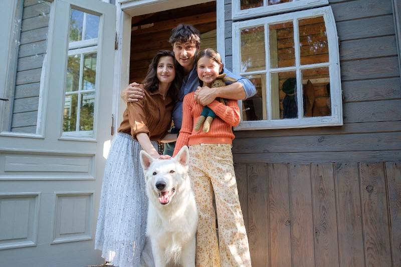 Uśmiechnięta rodzina składająca się z trzech osób i biały pies stoją w drzwiach drewnianego domu. Ojciec obejmuje matkę i córkę, która trzyma lalkę. Przez otwarte drzwi i okna wpadają promienie słońca.