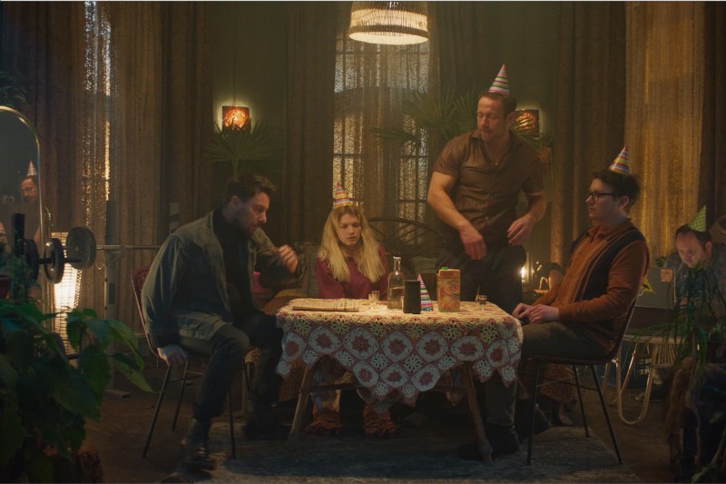 Czterech dorosłych w czapkach imprezowych siedzi przy stole z drinkami w słabo oświetlonym pokoju, udekorowanym roślinami i ciepłym oświetleniem. Wszyscy wydają się poważni lub zamyśleni, tworząc nieco ponurą atmosferę imprezy.