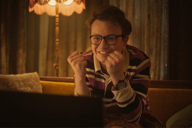 Młoda osoba w okularach i pasiastej koszuli siedzi na kanapie, uśmiecha się podekscytowana, trzymając obie pięści blisko twarzy, w ciepło oświetlonym pokoju, w tle pali się lampa.