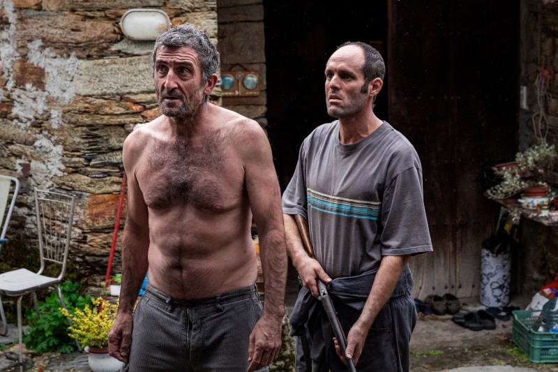 Dwóch mężczyzn w średnim wieku stoi na zewnątrz, w pobliżu rustykalnego kamiennego budynku; jeden jest bez koszuli i patrzy przed siebie, podczas gdy drugi, w pasiastej koszulce, trzyma strzelbę. Okolica wydaje się zagracona przedmiotami gospodarstwa domowego i roślinami.