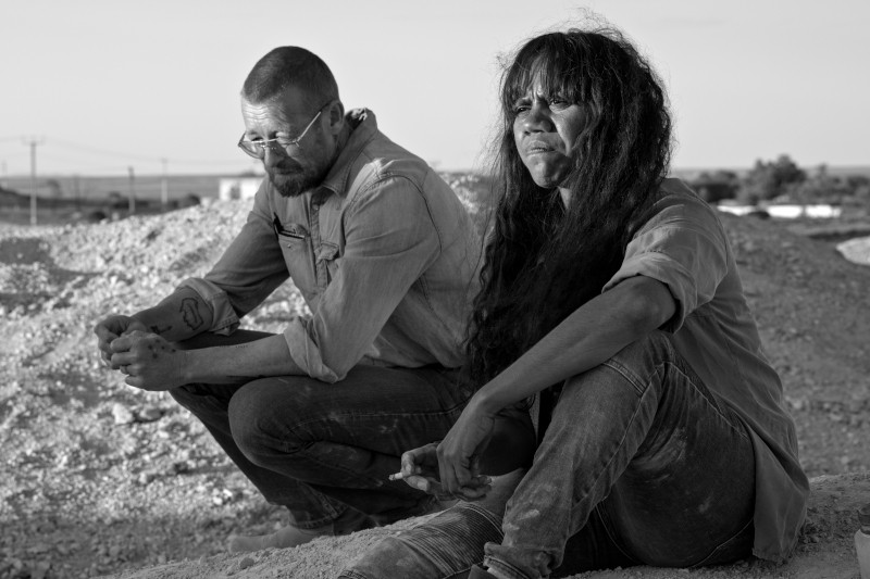 Dwie osoby siedzą na kamienistym gruncie na zewnątrz, wyglądając na zamyślonych i poważnych. Mężczyzna nosi okulary i ma krótkie włosy i brodę; kobieta ma długie ciemne włosy. Obraz jest czarno-biały.