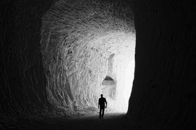 Przy wejściu do dużej jaskini o szorstkiej fakturze stoi sylwetka mężczyzny, a otwór jaskini przed nim jest oświetlony jasnym światłem.