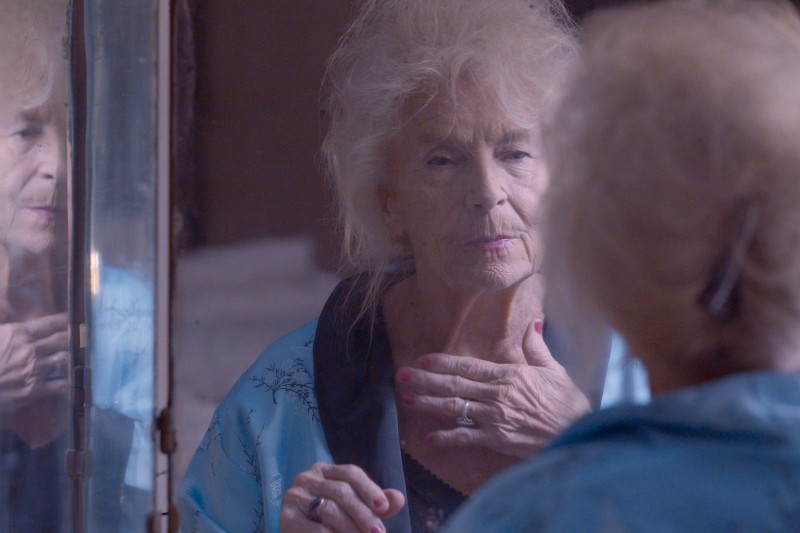 Starsza kobieta ubrana w niebieski szlafrok patrzy zamyślona na siebie w lustrze, delikatnie dotykając szyi jedną ręką. Jej odbicie jest widoczne w lustrze obok niej.