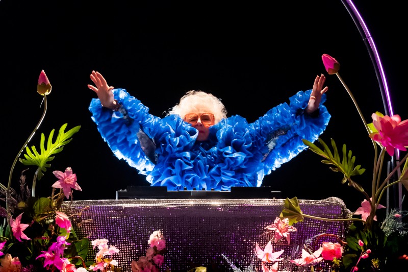 Osoba w jasnoniebieskim, falbaniastym stroju i pomarańczowych okularach stoi za błyszczącą powierzchnią, z uniesionymi ramionami, otoczona kolorowymi kwiatami na czarnym tle.