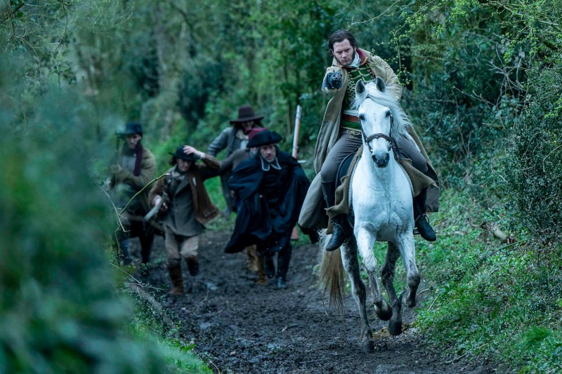 Mężczyzna na białym koniu wycelowuje pistolet do przodu, jadąc błotnistą leśną ścieżką, ścigany przez kilku mężczyzn w historycznych strojach i kapeluszach. Scena jest napięta i pełna akcji.