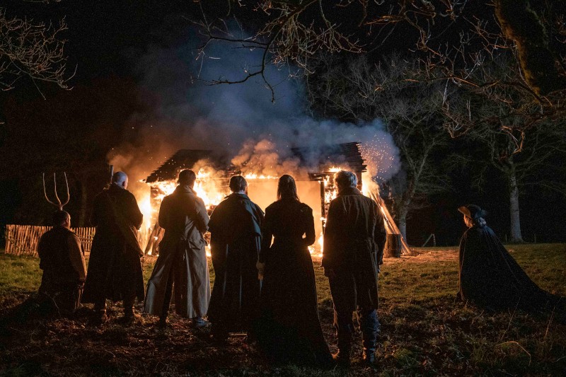Siedem osób ubranych w staromodne stroje stoi nocą w cieniu, patrząc na płonący drewniany dom, z którego wydobywają się płomienie i unoszący się dym, otoczony bezlistnymi drzewami.
