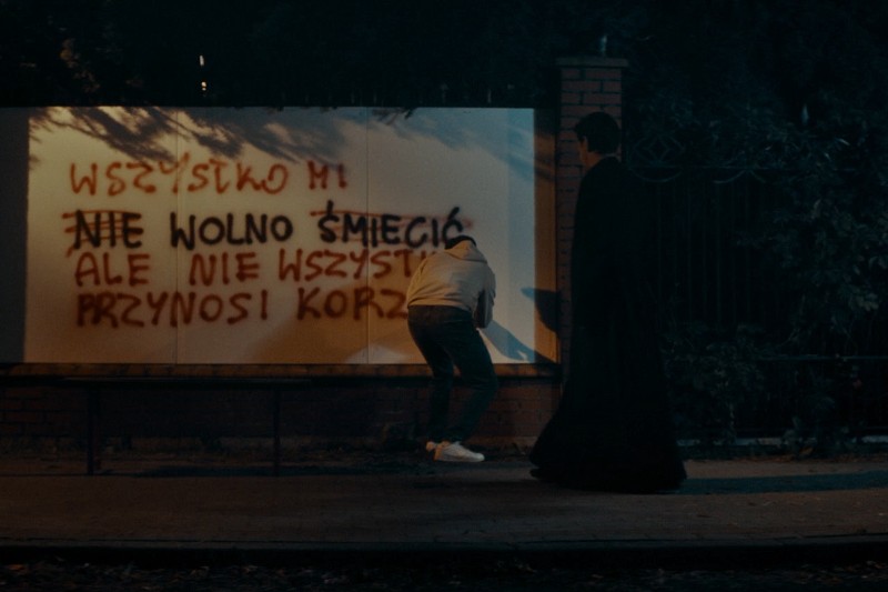 Osoba w bluzie z kapturem maluje sprayem polskie graffiti na ścianie nocą, podczas gdy inna osoba w ciemnym ubraniu stoi w pobliżu, częściowo w cieniu. W tle widać drzewa i płot.