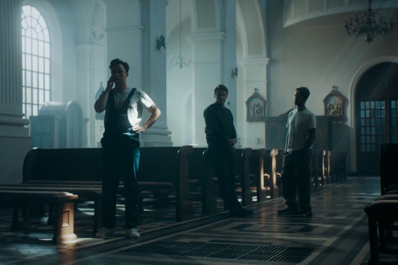 Trzech mężczyzn stoi w oświetlonym słońcem kościele; jeden rozmawia przez telefon, drugi stoi ze złożonymi dłońmi, a trzeci stoi swobodnie. Drewniane ławki i wysokie łukowate okna są widoczne w przestronnym, ozdobnym wnętrzu.