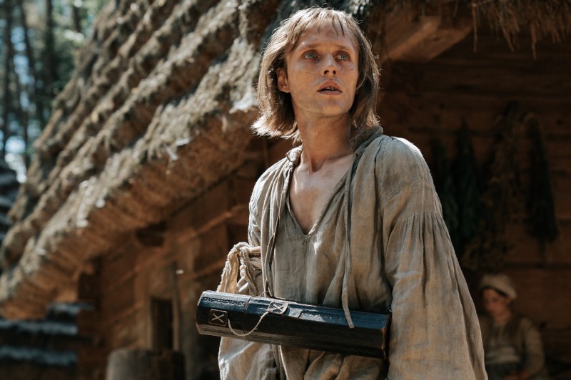 Osoba z włosami sięgającymi ramion, ubrana w staromodne, podarte ubranie, stoi przed rustykalnym budynkiem ze strzechą, trzymając czarne drewniane pudełko. Sceneria wydaje się historyczna lub średniowieczna.