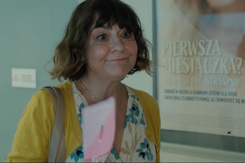 Kobieta z krótkimi brązowymi włosami, ubrana w żółty kardigan i kwiecistą bluzkę, uśmiecha się, trzymając różową teczkę. Na ścianie za nią widoczny jest rozmazany plakat z polskim tekstem.