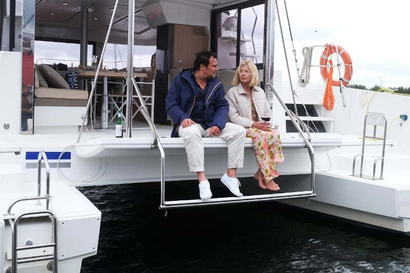 Mężczyzna i kobieta siedzą razem na tylnym pokładzie jachtu, rozmawiając i relaksując się. Kobieta trzyma kieliszek wina, a w pobliżu znajduje się otwarta butelka. Scena wychodzi na wodę i widać część wnętrza łodzi.